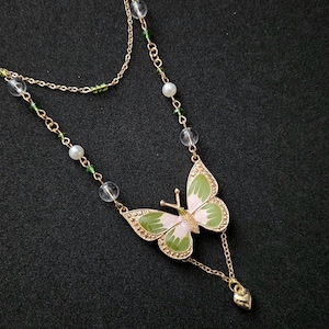 May include: A double-layered gold-tone chain necklace featuring a central butterfly pendant with green and white enamel wings. The chain is accented with small white pearl-like beads, clear spherical beads, and tiny green beads, ending in a small heart charm.