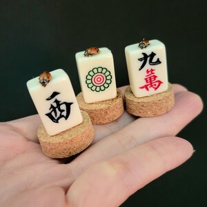 May include: Three ivory-colored Mahjong tiles, each with unique symbols and characters in black and red, are displayed on cork bases. A small ladybug rests on top of each tile. The tiles are held in a hand against a dark background.
