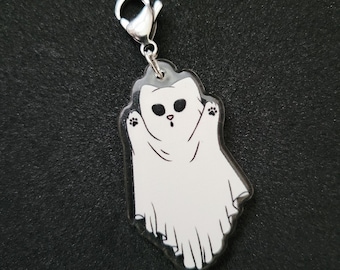 May include: A clear acrylic charm featuring a white cat wearing a ghost costume with black eyes, a small nose, and visible paw prints on the raised arms, attached to a silver-toned metal lobster claw clasp for hanging.