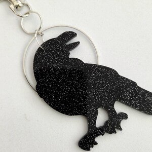 May include: A black raven keychain featuring a glittery finish and a silver-toned metal ring. The raven is depicted in profile with its beak open, attached to a circular silver hoop and a lobster claw clasp for easy attachment to bags or keys.
