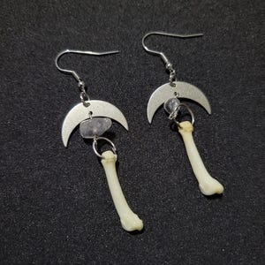 May include: A pair of unique earrings featuring a crescent moon charm, a small gemstone, and a bone-shaped pendant. The earrings have silver-colored ear wires and are displayed against a dark background.