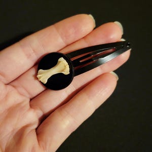 May include: Black metal hair clip with a small, round, black accent piece. The accent features a small, off-white bone. The hair clip is a simple, functional accessory.
