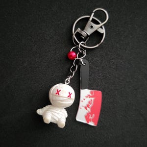 May include: A novelty keychain featuring a small white mummy figure with red cross eyes and a miniature cleaver with a red blood splatter design. The keychain includes a silver clasp and a red bead.