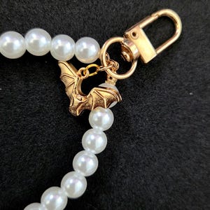 May include: A pearl necklace with a gold-colored bat charm and clasp. The necklace features large, round, white pearls. The bat charm is detailed with outstretched wings. The clasp is a simple, gold-colored lobster claw.