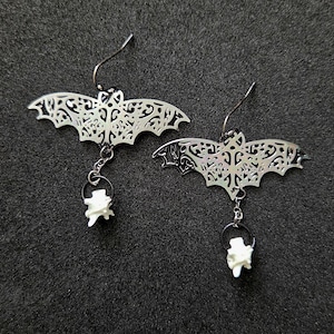 May include: A pair of silver-toned metal earrings featuring a filigree bat silhouette suspended from a hook. Each bat is attached to a small silver chain holding a single, white, textured bone-like vertebrae charm dangling at the bottom.