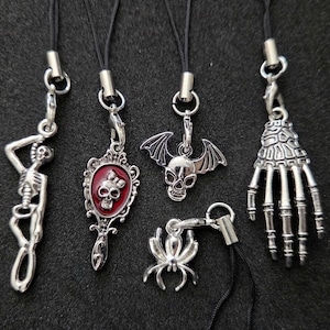 May include: Five silver-toned metal charms attached to black cords. The charms include a full skeleton, a hand mirror with a red inlay and skull, a winged skull, a spider, and a skeletal hand. Each charm features a lobster claw clasp.