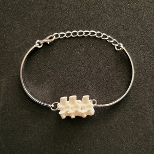 May include: A silver bracelet featuring three off-white vertebrae-shaped beads. The bracelet has an adjustable chain clasp. The background is a plain black.