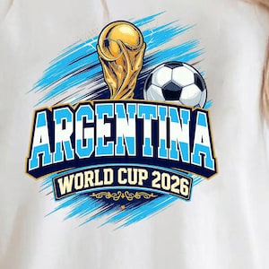 May include: White t-shirt featuring a graphic design for Argentina's World Cup 2026. The design includes a golden trophy, a soccer ball, and the word "ARGENTINA" in blue, with "WORLD CUP 2026" below. Blue brushstrokes complete the design.
