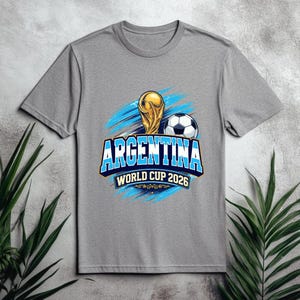 May include: A gray t-shirt featuring a graphic design for the Argentina World Cup 2026. The design includes a gold trophy, a soccer ball, and the text "ARGENTINA WORLD CUP 2026" in blue and white.
