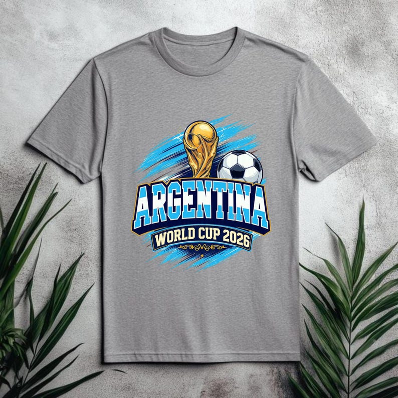 May include: A gray t-shirt featuring a graphic design for the Argentina World Cup 2026. The design includes a gold trophy, a soccer ball, and the text "ARGENTINA WORLD CUP 2026" in blue and white.