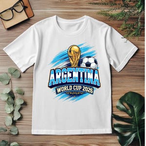 May include: White t-shirt featuring a gold trophy, soccer ball, and the text "ARGENTINA WORLD CUP 2026" in blue and white. The design includes blue brushstroke accents, perfect for soccer fans.