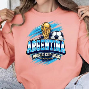 May include: A light pink sweatshirt featuring a graphic design for Argentina's World Cup 2026. The design includes a golden trophy, a soccer ball, and the word "ARGENTINA" in blue and white, with the text "WORLD CUP 2026".