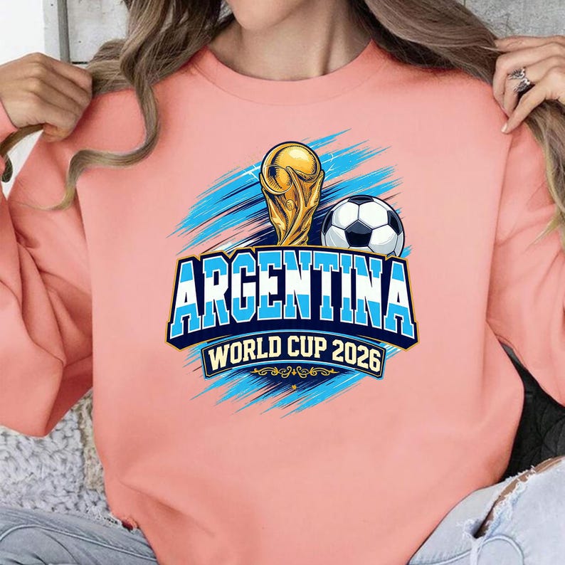 May include: A light pink sweatshirt featuring a graphic design for Argentina's World Cup 2026. The design includes a golden trophy, a soccer ball, and the word "ARGENTINA" in blue and white, with the text "WORLD CUP 2026".
