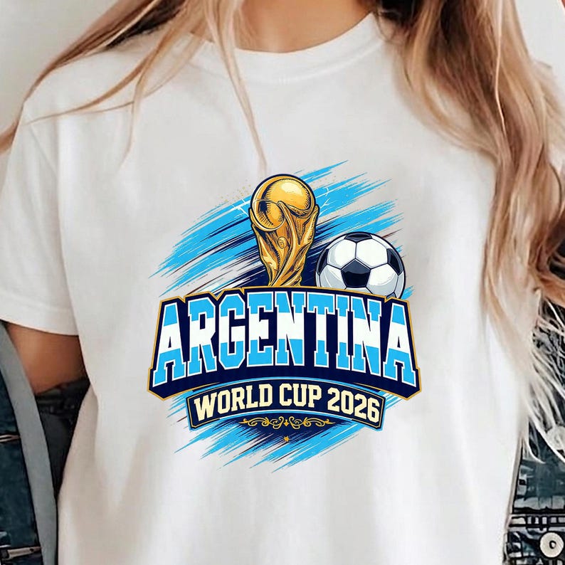 May include: White t-shirt featuring a graphic design for Argentina's World Cup 2026. The design includes a golden trophy, a soccer ball, and the word "ARGENTINA" in blue, with "WORLD CUP 2026" below. Blue brushstrokes complete the design.