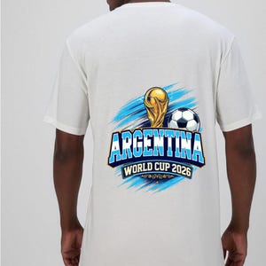 May include: White t-shirt featuring a graphic design for Argentina's World Cup 2026. The design includes a gold trophy, a soccer ball, and the word "ARGENTINA" in blue and white, with the text "WORLD CUP 2026" below.