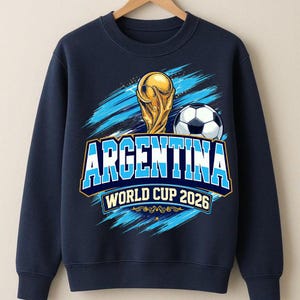 May include: Navy blue sweatshirt featuring a gold trophy and a soccer ball. The word "ARGENTINA" is displayed in a bold, blue and white font, with "WORLD CUP 2026" below. The design includes blue brushstroke accents.