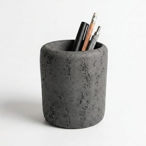 May include: A cylindrical, gray concrete desk organizer with a textured surface. The organizer holds a black pen, a pencil, and a silver mechanical pencil. The container has a rounded top and is set against a white background.