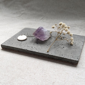 May include: A gray rectangular stone tray with a brown edge, featuring a small silver pendant necklace, a purple amethyst crystal, and a sprig of white flowers. The items are arranged on a light gray fabric background.