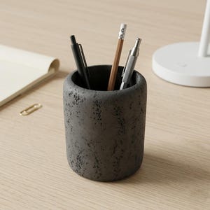Op de afbeelding: Een donkergrijze, getextureerde pennenhouder met een cilindrische vorm. De houder bevat een zwarte pen, een zilveren pen en een houten potlood. Een notitieboek en een gouden paperclip liggen op het lichte houten oppervlak.