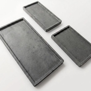 Könnte beinhalten: Drei rechteckige, graue Tabletts unterschiedlicher Größe sind auf einer weißen Oberfläche angeordnet. Die Tabletts haben einen erhöhten Rand und eine strukturierte Oberfläche. Die Tabletts sind wahrscheinlich aus Stein oder Beton.