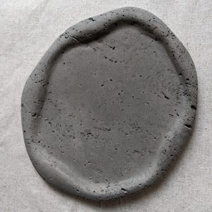 Puede incluir: Una bandeja de hormigón gris de forma irregular con un borde ligeramente elevado. La superficie tiene un aspecto texturizado con pequeños agujeros e imperfecciones. La bandeja está diseñada para uso decorativo o funcional.