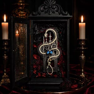 May include: A black wooden display case containing a coiled white snake skeleton and a blue butterfly. The interior features dark foliage and architectural elements. Two brass candlesticks with lit white candles stand on either side of the case.