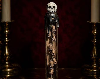 May include: A tall, clear glass test tube containing a vertical arrangement of small, dried, brown skull-shaped objects. The top is sealed with a cork, black wax drippings, and a small, detailed white skull figurine perched on the very top.