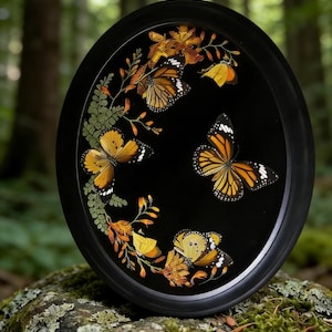 May include: A black, oval-shaped decorative tray featuring several orange and black butterflies with white spots. The butterflies are arranged on a black background with dried leaves and greenery, all within a black frame.