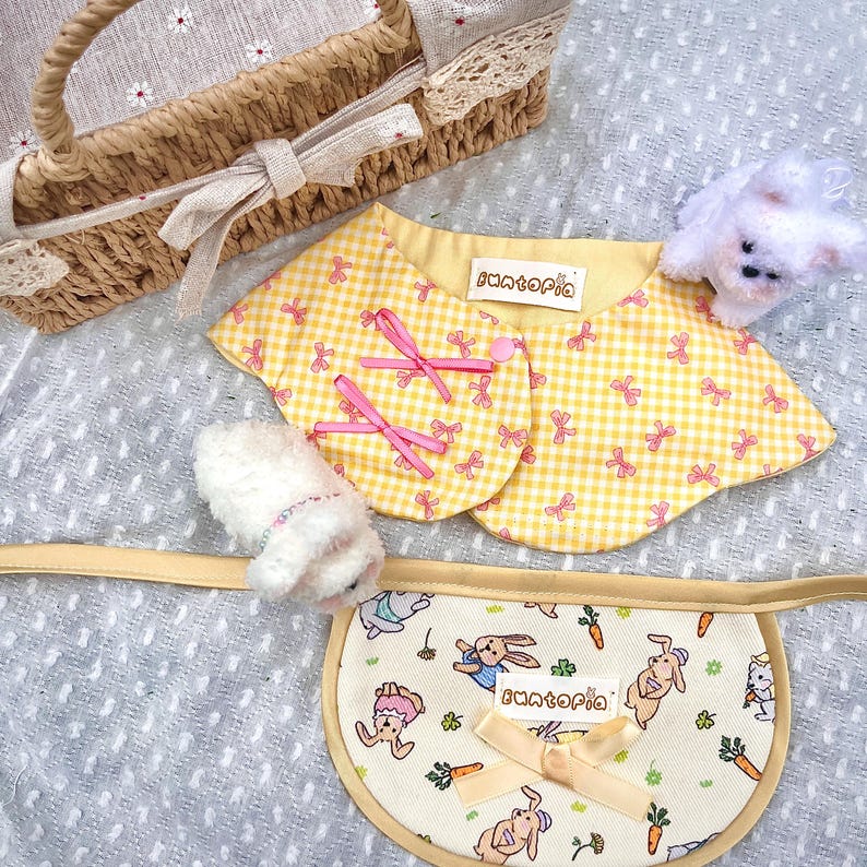 May include: A yellow and white gingham fabric collar with pink bows and the text "Funtopia" on a white label. The collar is on a gray surface with a white stuffed animal and a white wicker basket with a brown handle.