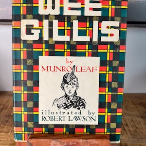 May include: A hardcover book titled Wee Gillis by Munro Leaf, illustrated by Robert Lawson. The cover features a green, red, and yellow plaid pattern with white text. A central cream-colored inset displays a black and white illustration of a person wearing a tam-o'-shanter hat and a kilt, with the author and illustrator names printed in black and red.