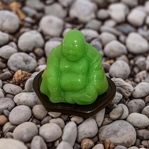 Puede incluir: Una pequeña figura de jade verde de un Buda sonriente sentado en una postura meditativa sobre una base de madera negra. La figura está rodeada de pequeñas piedras grises.