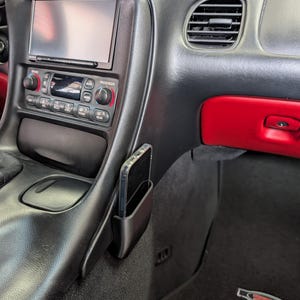 May include: Black car interior with a smartphone in a black phone holder. The dashboard features a digital display, radio controls, and air vents. The interior has red and black accents.
