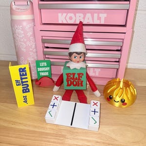 May include: A pink Kobalt tool chest, a pink water bottle with white floral patterns, and an Elf on the Shelf doll wearing a green box labeled Elf Doh. Includes a yellow Elf Butter sign, a green Lets Squishy Trade sign, a white game piece, and a gold dumpling toy.