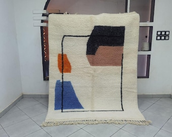 May include: Cream-colored, rectangular rug with an abstract design. The design features a black outline with geometric shapes in orange, blue, black, and brown. The rug has a fringe along the bottom edge and is displayed against a white wall.