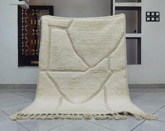 May include: Cream-colored, geometric patterned rug with a textured surface. The rug features a design of irregular shapes and lines, with a fringe along the bottom edge. The rug is displayed indoors.