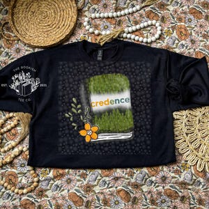 May include: Black crew neck sweatshirt with a graphic of a book with the word "credence" on the cover. The book design features a green grassy top and a forest scene. The sweatshirt also has the text "The Bookish Tee Co. Est. 2021" on the left sleeve.