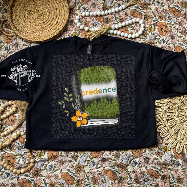 May include: Black crew neck sweatshirt with a graphic of a book with the word "credence" on the cover. The book design features a green grassy top and a forest scene. The sweatshirt also has the text "The Bookish Tee Co. Est. 2021" on the left sleeve.