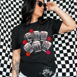 May include: Black t-shirt featuring a graphic of book covers with titles like "Corrupt" and "Nightfall," surrounded by red roses. The text "THE BOOKISH TEE" and the year 2021 are printed at the bottom. The shirt is a casual wear item.