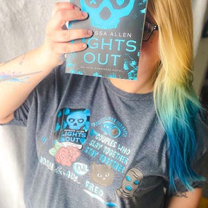 May include: A person holding a book titled "Lights Out" by Tessa Allen, with a skull design. The person is wearing a gray t-shirt with a matching graphic. The shirt features the text "Couples Who Slay Together, Stay Together."