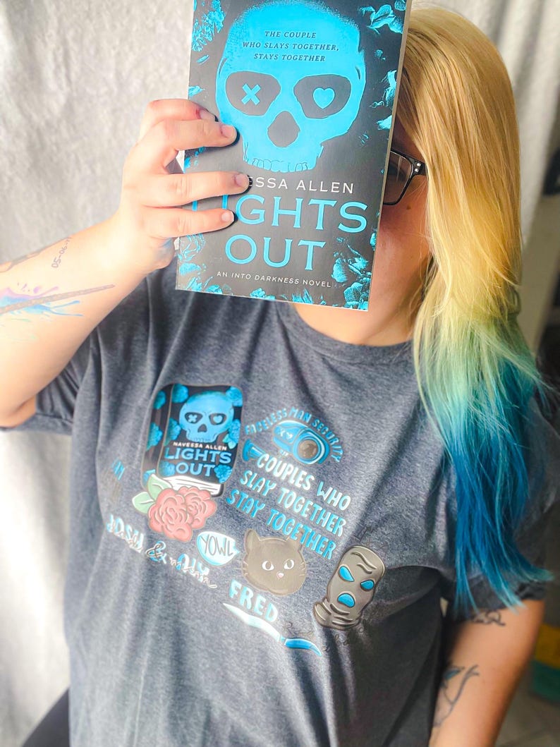 May include: A person holding a book titled "Lights Out" by Tessa Allen, with a skull design. The person is wearing a gray t-shirt with a matching graphic. The shirt features the text "Couples Who Slay Together, Stay Together."