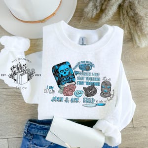 May include: White crewneck sweatshirt with various graphic designs in blue, black, and pink. The graphics include a skull, a cat, a balaclava, and the text "LIGHTS OUT", "COUPLES WHO SLAY TOGETHER STAY TOGETHER", and "I AM BATMAN".