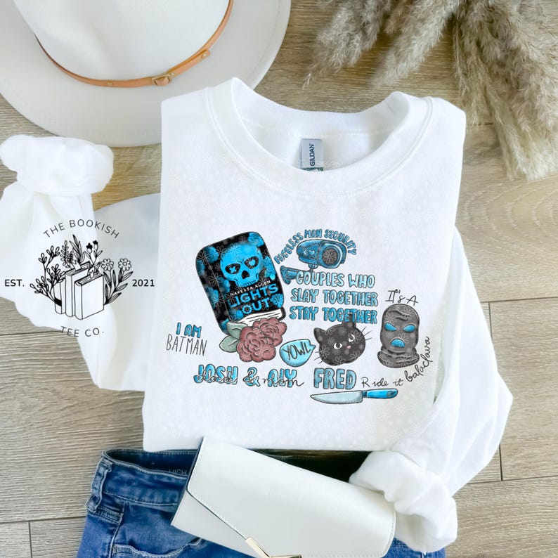 May include: White crewneck sweatshirt with various graphic designs in blue, black, and pink. The graphics include a skull, a cat, a balaclava, and the text "LIGHTS OUT", "COUPLES WHO SLAY TOGETHER STAY TOGETHER", and "I AM BATMAN".