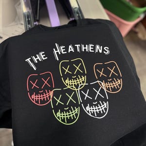 Może przedstawiać: Czarny T-shirt z napisem "THE HEATHENS" w białych literach. Na koszulce nadrukowane są cztery neonowe, zielone, r&oacute;żowe, pomarańczowe i białe kontury uśmiechniętych twarzy z X zamiast oczu.