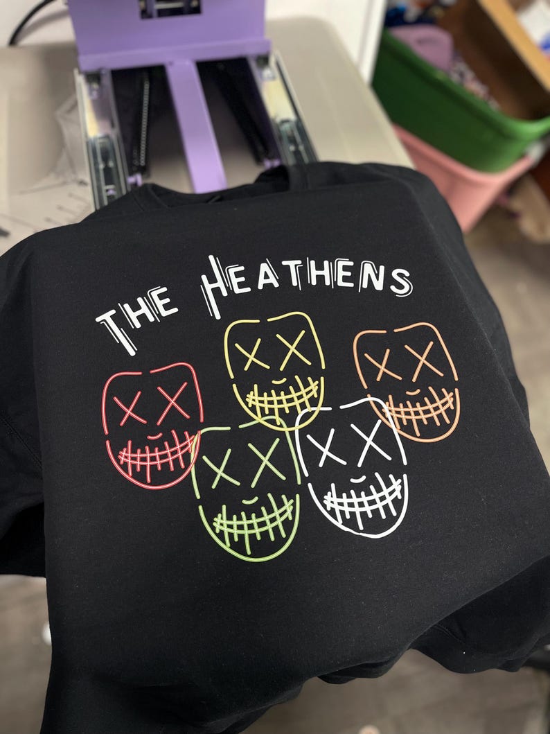 Może przedstawiać: Czarny T-shirt z napisem "THE HEATHENS" w białych literach. Na koszulce nadrukowane są cztery neonowe, zielone, r&oacute;żowe, pomarańczowe i białe kontury uśmiechniętych twarzy z X zamiast oczu.