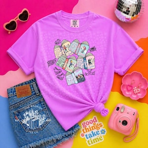 May include: A lilac Comfort Colors t-shirt featuring a graphic of illustrated books with people and the text "Briary", "Deal", and "Score". The shirt is paired with denim shorts, sunglasses, and a pink camera. A sticker reads "good things take time".