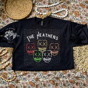 Może przedstawiać: Czarna bluza z białym nadrukiem graficznym z tekstem "The Heathens" i pięcioma stylizowanymi twarzami z oznaczeniami "XX". Grafika jest otoczona wzorem małych białych kropek.