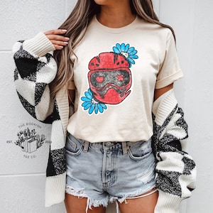 K&ouml;nnte beinhalten: Beiges T-Shirt mit roter Helm-Grafik, herzf&ouml;rmigen Augen und einem Kreuz, umgeben von blauen Blumen. Das T-Shirt wird mit Jeansshorts und einer schwarz-wei&szlig; karierten Strickjacke getragen. Das Logo von The Bookish Tee Co. ist sichtbar.