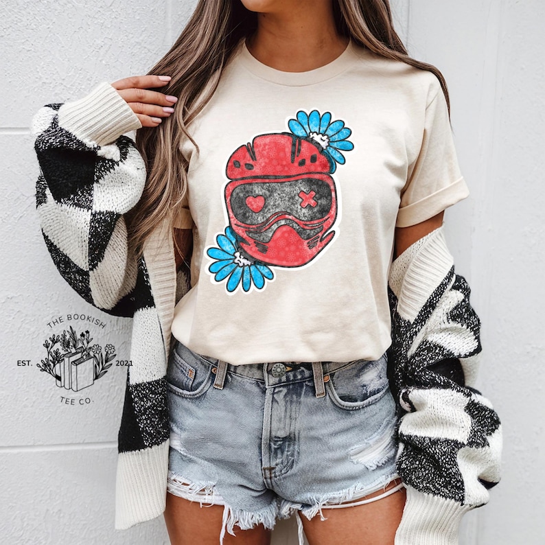 K&ouml;nnte beinhalten: Beiges T-Shirt mit roter Helm-Grafik, herzf&ouml;rmigen Augen und einem Kreuz, umgeben von blauen Blumen. Das T-Shirt wird mit Jeansshorts und einer schwarz-wei&szlig; karierten Strickjacke getragen. Das Logo von The Bookish Tee Co. ist sichtbar.