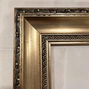 May include: A gold picture frame with a decorative ornate design. The frame has a raised edge and a smooth, polished finish.