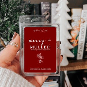 May include: A red wax melt package with the words "merry + MULLED" in white script. The scent is cranberry, apple, and marmalade. The package is 2.6 oz and contains beeswax and tallow melts. The package is held in a person's hand.